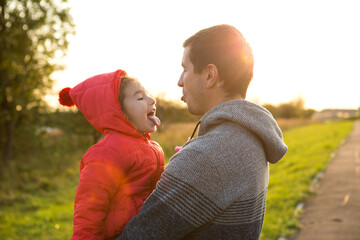 Little girl in red jacket with a hood hugs and stick out tongue her dad, smiles. Happy family, children's emotions, father's day, bright rays of the sun, Caucasian appearance. Space for text.