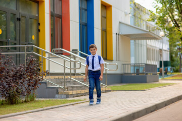 happy schoolboy in white shirt, blue tie and backpack left school with multicolored windows