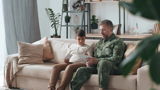 Medium shot of male army veteran in military uniform sitting on couch in living room and showing photos to his son while sharing deployment stories