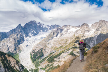 Fototapeta na wymiar Woman with backpack walking on a trail at high altitude, in Italian alps. High peaks with glaciers and clouded sky in the background