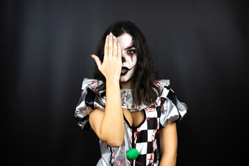 woman in a halloween clown costume over isolated black background covering one eye with hand, confident smile on face and surprise emotion.