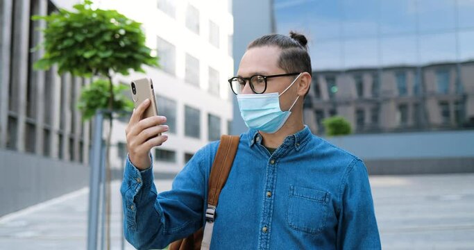 Caucasian young man in glasses and medical mask talking via webcam on mobile phone at steet. Handsome male in eyeglasses videochatting the cellphone outdoor. Video call conversation. Videochat.