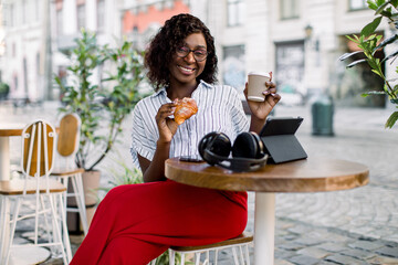 Pretty African business lady having lunch time outdoor in city cafe. African American smiling woman sitting at the table in city cafe, holding a cup of coffee and croissant, looking at camera