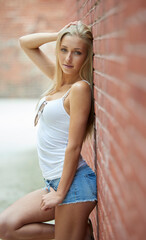 Beautiful young blonde woman poses against brick wall wearing white tank top and denim shorts