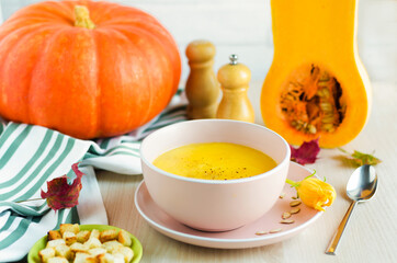 Foliage leaves, flowers and creamy pumpkin soup with vegetables, seeds and croutons in a bowl on light wooden background. Thanksgiving Day. traditional autumn dish, menu on the table.