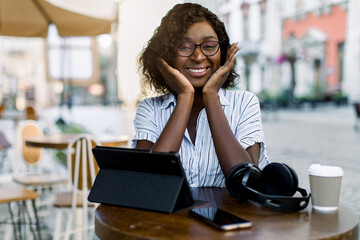 Pretty black business lady , sitting at the table in outdoor city coffee shop, with headphones, smartphone and tablet ipad, enjoying her lunch time outside office, smiling to camera