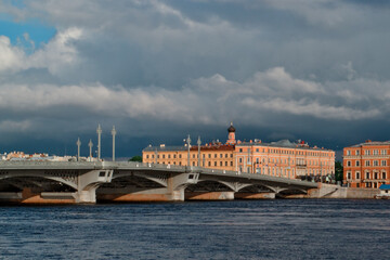 Fototapeta premium Saint Petersburg's big Blagoveshchensky Bridge over blue Neva River leading to embankment with colorful bright red and orange historical buildings on shore in light of setting sun. Dark storm clouds