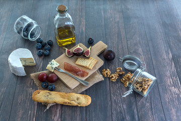 Slate dish with cheese, figs, bread, ham, blueberries and nuts on a wooden surface.