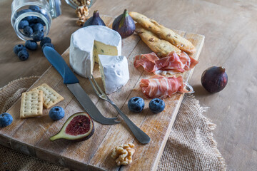 Wooden board with cheese, figs, bread, ham, blueberries and nuts.