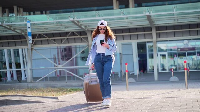 Traveling hipster girl student tourist walking with suitcase and smartphone in hands uses application to order a taxi. Young in casual clothes woman walks on urban city background enjoys mobile phone