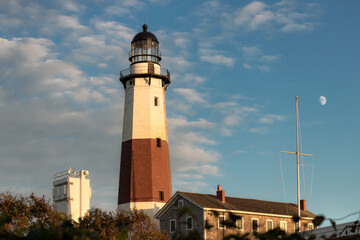 Fototapeta na wymiar Golden sunset light hitting a tall stone lighthouse, with a moon rising in the background. Montauk Point, New York 