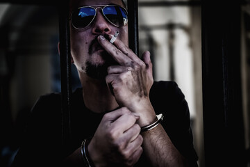 Arrested and handcuffed man imprisoned for crime smokes from behind bars. Gangster and pain concept.