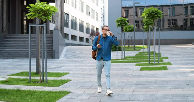 Caucasian young man walking steps downn at street, tapping on smartphone and sipping drink. Male gooing down the ladder outdoor, texting mesage on phone and drinking coffee to-go. Rushing in morning.