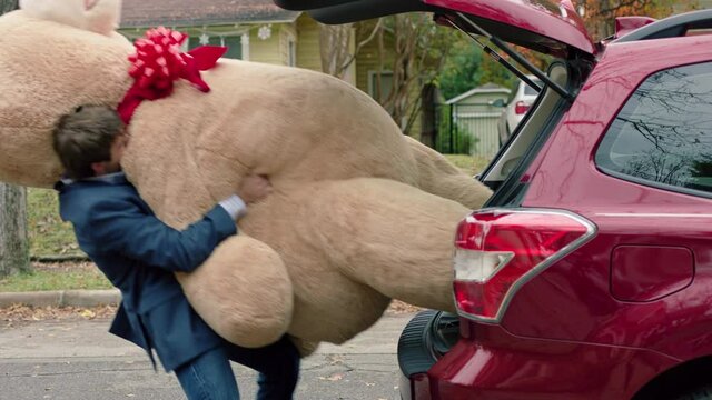 MCU of a man fighting to get a large valentines day teddy bear out of the trunk of his car.