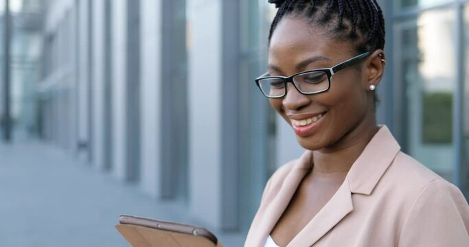 Close up of African American young smiled businesswoman in glasses tapping and scrolling on tablet device. Woman texting message on computer outdoors. Confident female using gadget and chatting.