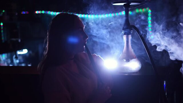 Close up side view of a girl smoking hookah on blue neon background. Media. Female while smoking a hookah in a club, exhaling a fog cloud and chilling.