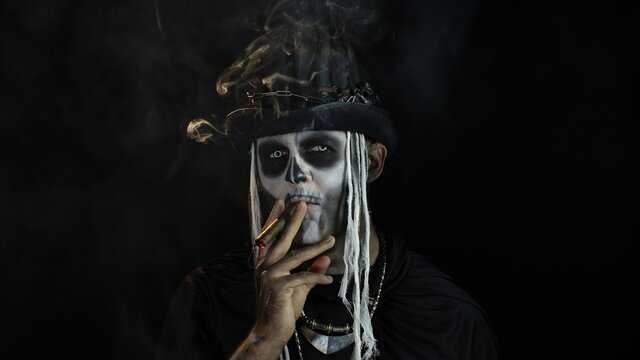 Sinister man with horrible Halloween skeleton makeup smoking cigar, making faces