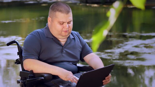 wheelchair man. Handicapped man. young disabled man, sitting in an automated wheelchair and working on a laptop, in a city park, by the lake, on autumn sunny day.