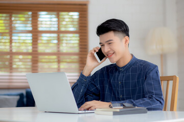 Young asian man talking phone and work from home with laptop computer, freelance using notebook to internet online and speak on smartphone, domestic life, business and communication concept.