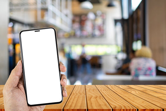Mockup image of hand holding smartphone with top wooden table in blurred coffee shop background.