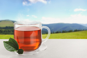 Glass cup of fresh hot tea on wooden table against blurred mountain landscape. Space for text