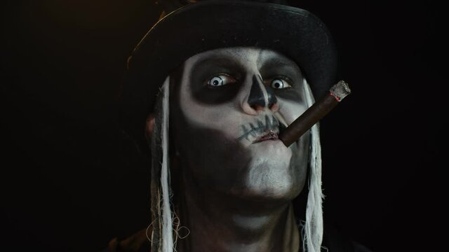 Sinister man with horrible Halloween skeleton makeup smoking cigar, making faces, smiling, laughing
