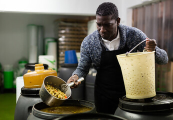 Afro-american man takes olives from barrel with colander in food factory