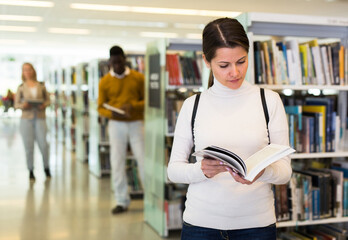 Adult attractive woman reading book while standing near bookshelves in public library. High quality...