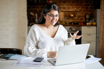 Cheerful plump young female economist wearing glasses and wireless earphones sitting at desk with...