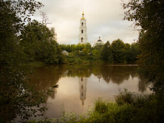 The high white bell tower and a dome of a Russian Orthodox cathedral are reflecting in still water of a small pond. The picture is framed by green foliage. Overcast sky, no people.