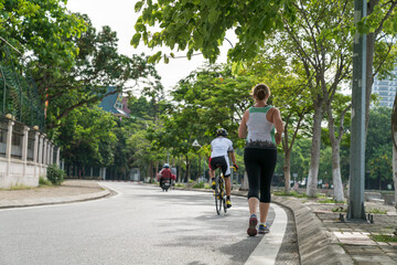 Sporty Caucasian woman running on the road in the Asian city park with green tree row in early morning