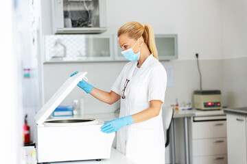 Three quarter length of attractive blond female lab assistant in sterile uniform with rubber gloves na d surgical mask on closing machine for blood samples while standing in laboratory.