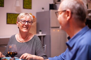 Elderly woman wearing glasses while having festive dinner with husband. Happy cheerful senior elderly couple dining together in the cozy kitchen, enjoying the meal, celebrating their anniversary.