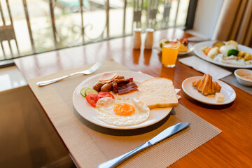 Fresh breakfast table next to window with bread, pastry, egg, fruit, juice, coffee cup