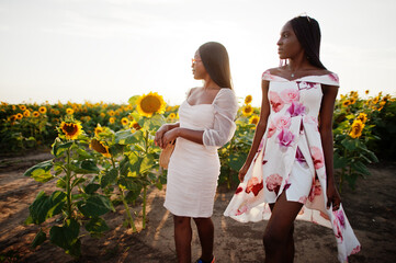 Two pretty young black friends woman wear summer dress pose in a sunflower field.