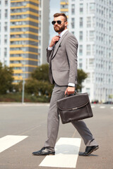 side view on caucasian businessman in suit or tuxedo walking with bag in hands, wearing sunglasses. in city streets