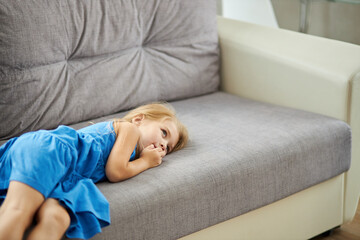 Obraz na płótnie Canvas sad caucasian little girl lying on sofa alone, wearing domestic dress, depressed upset girl looking away