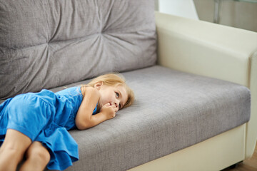 sad caucasian little girl lying on sofa alone, wearing domestic dress, depressed upset girl looking away