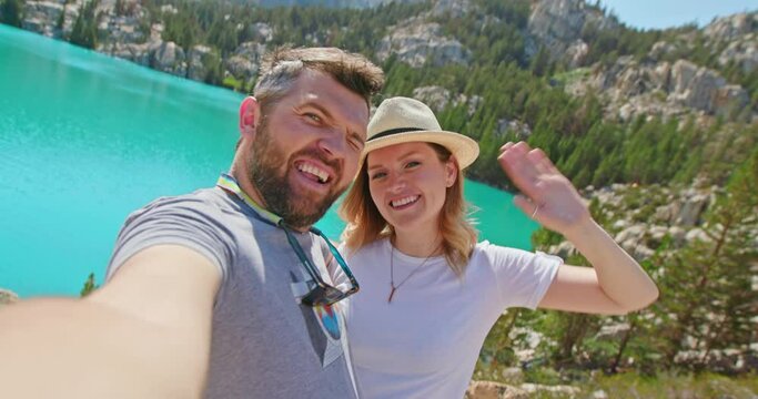 Traveling couple takes selfie picture or video on peak of mountain with beautiful blue lake on background at sunny summer day. Young romantic couple posing in outdoor, USA. 4K happy people travel shot