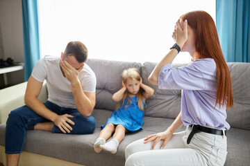 sad, desperate little girl during parents quarrel, she clog the ears sitting on sofa at home, angry parents fighting. worried upset small daughter hurt by fathers and mothers break up or divorce