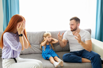 sad, desperate little girl during parents quarrel, she clog the ears sitting on sofa at home, angry parents fighting. worried upset small daughter hurt by fathers and mothers break up or divorce