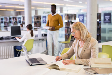 Fototapeta na wymiar Mature confident woman with laptop and book in public library. High quality photo