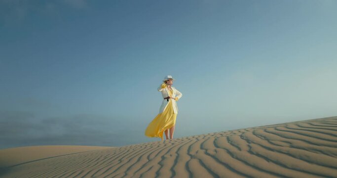 Beautiful fashion footage. Woman in stylish safari outfit is on wavy textured sand desert surface. Slow motion fashion shot with blue sky on background. Advertisement cinematic view of stylish woman