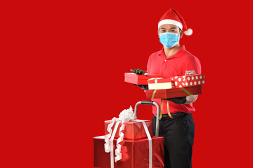 Happy young Asian delivery man in red uniform, medical face mask, protective gloves, christmas hat carry boxes of presents in hands on red background during COVID-19 outbreak and christmas festivities