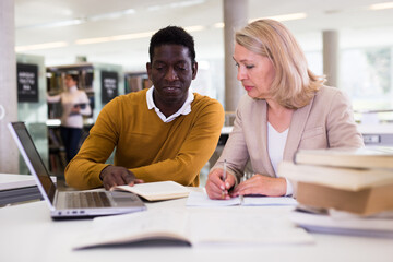 Friendly female tutor helping to diligent african-american man preparing for exam in library. High quality photo