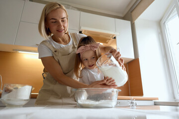 happy mother baking with little daughter in apron and cook hat working with flour , bowl and spoon preparing dough teaching the kid baking and having fun together.