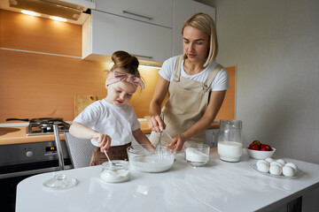happy mother baking with little daughter in apron and cook hat working with flour , bowl and spoon preparing dough teaching the kid baking and having fun together.