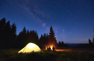 Night camping near fire, forest and mountains on background. Group of friends warming up near bright bonfire. People sitting near tourist illuminated tent under night sky full of stars and milky way. © anatoliy_gleb