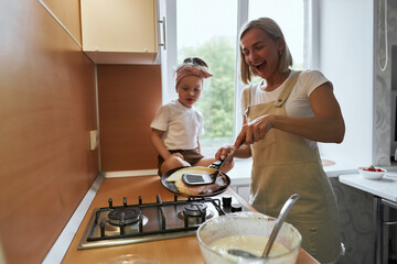 happy mother baking with little daughter in apron and cook hat working with flour , bowl and spoon preparing dough teaching the kid baking and having fun together.