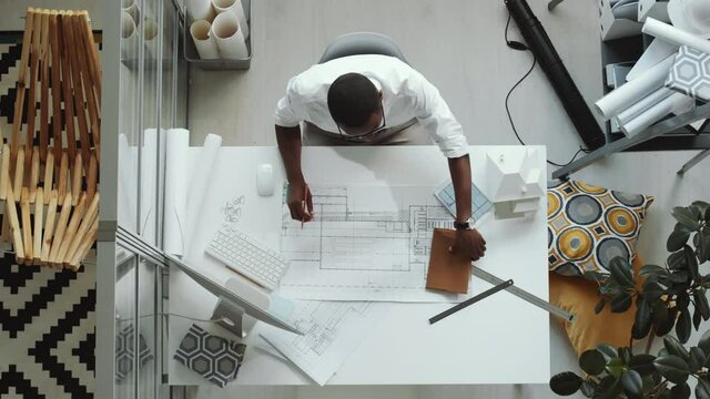 Top down shot of African American architect using ruler, writing down measurements in notepad and drawing floor plan with pencil on paper while working at office desk